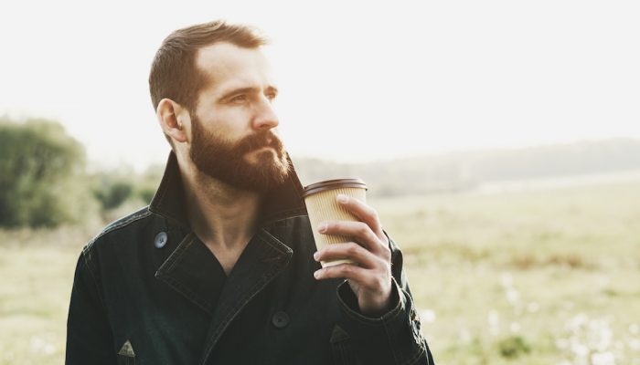 handsome bearded man with paper cup of morning coffee walking in park